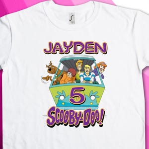 Pode incluir: Camiseta branca com um gráfico de Scooby-Doo roxo e verde. O gráfico apresenta a Mystery Machine com os personagens do programa. O texto "Jayden 5 Scooby-Doo!" está impresso na camiseta.