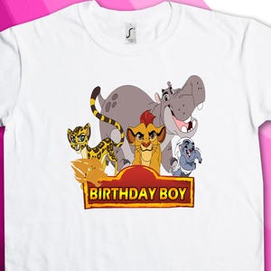 Puede incluir: Camiseta blanca con un gráfico de personajes de la caricatura de El Rey León. Los personajes incluyen Simba, Nala, Pumbaa y Timon. El gráfico está rodeado de una pancarta roja con el texto "Birthday Boy".
