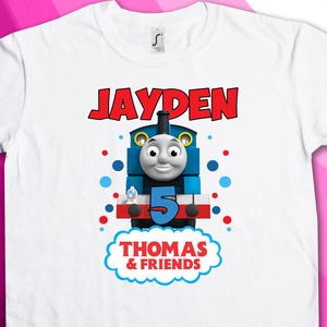 Puede incluir: Camiseta blanca con un diseño de Thomas la locomotora azul y rojo. El texto "JAYDEN" está impreso en rojo sobre el tren. El texto "THOMAS & FRIENDS" está impreso en rojo debajo del tren.