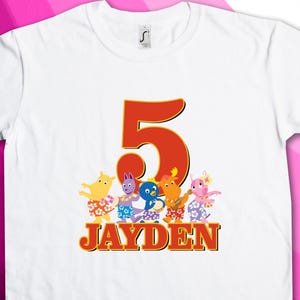 May include: White t-shirt featuring a large orange number 5 and the name "JAYDEN" in orange and yellow. Below the number are cartoon characters in colorful outfits. The shirt is a birthday-themed item.