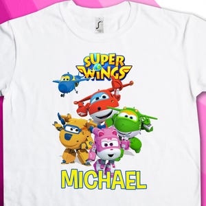 May include: White t-shirt featuring a colorful graphic of the Super Wings characters, including airplanes and robots. The name "MICHAEL" is printed in yellow block letters below the characters. The shirt has a crew neck and short sleeves.
