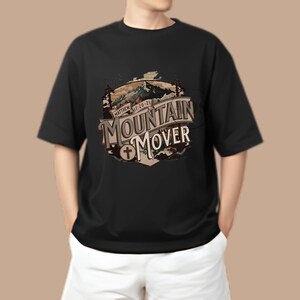 Peut inclure: T-shirt noir avec un motif graphique représentant une chaîne de montagnes et le texte "Mountain Mover" avec une croix. Le texte "Matthew 17:20-21" est également inclus dans le motif.
