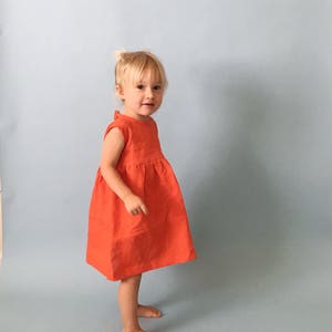 K&ouml;nnte beinhalten: Ein junges M&auml;dchen in einem leuchtend orangefarbenen, &auml;rmellosen Kleid steht vor einem hellblauen Hintergrund.