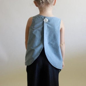 May include: A young person wearing a light blue sleeveless top with a single white button on the back. The top has a unique curved shape in the back. The person is also wearing black pants.