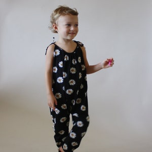May include: A young child wearing a black and white floral jumpsuit. The jumpsuit has a scoop neck and thin straps. The child is standing on a white background.