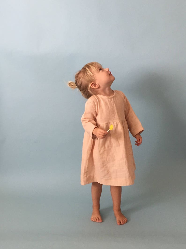 K&ouml;nnte beinhalten: Ein junges M&auml;dchen tr&auml;gt ein hellrosa Kleid mit langen &Auml;rmeln und einem gerafften Rock. Sie h&auml;lt einen gelben Lutscher in der Hand und schaut nach oben.