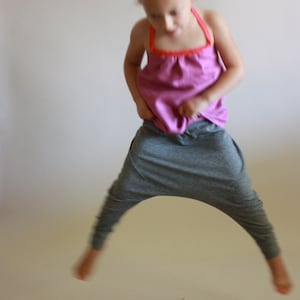 May include: A young girl wearing a pink tank top and grey striped trousers jumps in the air with her arms at her sides.