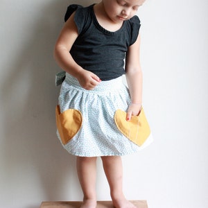 Puede incluir: Una niña pequeña con una camisa negra y una falda blanca con bolsillos en forma de corazón amarillos. La falda tiene un estampado de lunares blancos y azules.