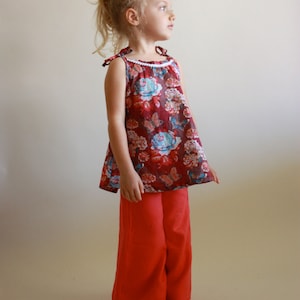 Könnte beinhalten: Ein junges Mädchen trägt ein rotes, ärmelloses Oberteil mit Blumenmuster und eine rote Hose. Das Oberteil hat einen weißen Saum am Ausschnitt.