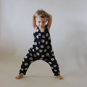Op de afbeelding: Een jong kind draagt een zwart-witte jumpsuit met een madeliefjesprint. De jumpsuit heeft een ronde hals en dunne bandjes. Het kind staat met zijn armen boven zijn hoofd.