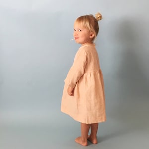 K&ouml;nnte beinhalten: Ein junges M&auml;dchen tr&auml;gt ein hellrosa Kleid mit langen &Auml;rmeln und einem gerafften Rock. Sie steht vor einem hellblauen Hintergrund.