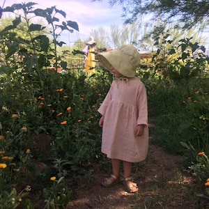 K&ouml;nnte beinhalten: Ein kleines Kind in einem rosa Leinenkleid und einem hellgr&uuml;nen Sonnenhut steht in einem Garten mit gelben Blumen.