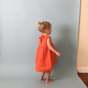K&ouml;nnte beinhalten: Ein kleines Kind tr&auml;gt ein helloranges, &auml;rmelloses Kleid mit Schleife am R&uuml;cken. Das Kind steht auf einem hellblauen Hintergrund.