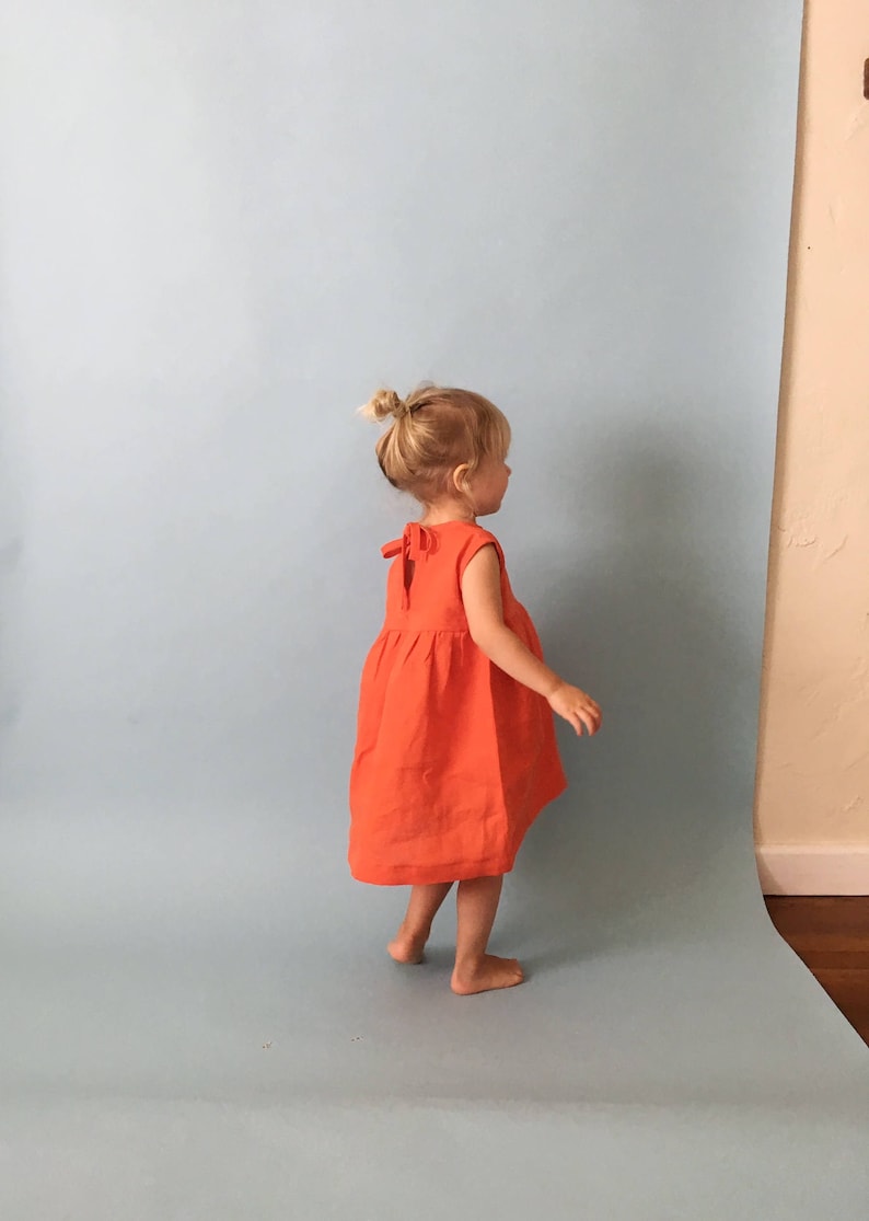 K&ouml;nnte beinhalten: Ein kleines Kind tr&auml;gt ein helloranges, &auml;rmelloses Kleid mit Schleife am R&uuml;cken. Das Kind steht auf einem hellblauen Hintergrund.