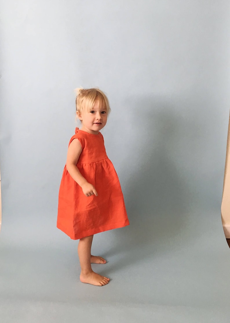 K&ouml;nnte beinhalten: Ein junges M&auml;dchen in einem leuchtend orangefarbenen, &auml;rmellosen Kleid steht vor einem hellblauen Hintergrund.
