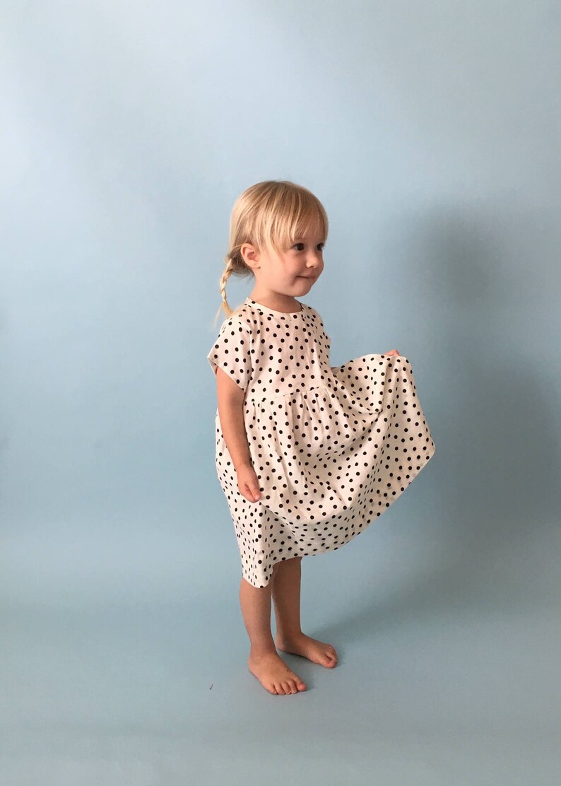 K&ouml;nnte beinhalten: Ein junges M&auml;dchen tr&auml;gt ein wei&szlig;es Kleid mit schwarzen Punkten. Das Kleid hat einen gerafften Rock und kurze &Auml;rmel. Das M&auml;dchen steht auf einem hellblauen Hintergrund.