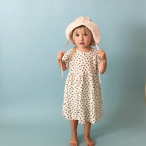 K&ouml;nnte beinhalten: Ein kleines Kind tr&auml;gt ein wei&szlig;es Kleid mit schwarzen Punkten und einen rosa Sonnenhut. Das Kind steht auf einem hellblauen Hintergrund.