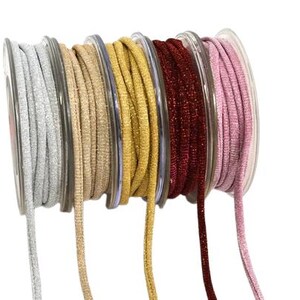 May include: Five spools of thin, round elastic cord arranged in a row. The cords feature a metallic, glittery finish in silver, beige, gold, deep red, and light pink. Each cord is wound around a clear plastic spool with a visible center core.