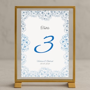 Op de afbeelding: Een goudkleurige tafelnummerkaart met een blauwe bloemenrand. Het nummer "3" staat in blauw, met het woord "Table" erboven. De namen "Victoria & Gabriel" en de datum "05.05.2025" staan ook op de kaart.