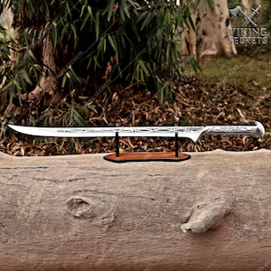 Op de afbeelding: Een zilverkleurig metalen zwaard met een ingewikkeld geëtst patroon langs het lemmet en het handvat. Het zwaard is horizontaal gemonteerd op een zwarte standaard met een houten voet, met een slanke punt en een afgerond pommelontwerp.