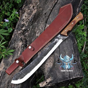 Op de afbeelding: Een grote, gebogen machete met een houten handvat en een leren schede. Het lemmet is zilverkleurig en het handvat bruin. De schede is van bruin leer met witte stiksels. De machete ligt op een houten oppervlak.