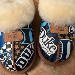 May include: A pair of blue and white UGG slippers with a Duke University theme. The slippers are decorated with a variety of Duke logos and designs, including the Duke Blue Devils mascot, the Duke University logo, and the words "Duke" and "University".
