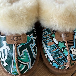 May include: A pair of brown UGG slippers with a white fur lining. The slippers are decorated with a green, blue, and white design featuring the words "Tulane" and "Green Wave".