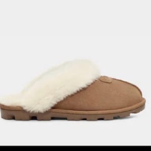 May include: A brown suede slipper with a cream-colored shearling lining and trim. The slipper has a rubber sole and a rounded toe. The design is a slip-on style, perfect for indoor wear.