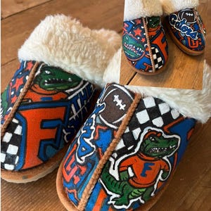 Peut inclure: Une paire de pantoufles moelleuses, bleues, orange et blanches sur le thème des Florida Gators. Les pantoufles présentent un mascotte de gator, la lettre F et un ballon de football.
