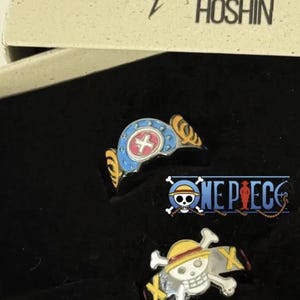 May include: Two metal rings featuring One Piece anime designs inside a black display box labelled Craft Hoshin. One ring is blue with a red and white cross emblem, while the other is a silver skull and crossbones with a yellow straw hat.