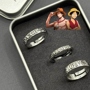 May include: Three silver-toned metal rings with open-ended bands, each featuring engraved text. One ring displays the word "LUFFY" with a globe icon, another reads "DEATH," and the third features "ACE" with a cross symbol, presented in a metal case.