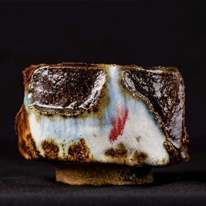 Peut inclure: Un bol à thé en céramique avec un extérieur brun foncé texturé et une finition brillante. La surface présente un motif de coulure d'émail blanc s'étendant verticalement, accentué par une petite marque rouge vive près du centre.