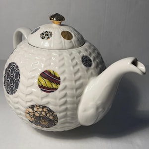 May include: White ceramic teapot with a textured surface and colorful, patterned circles. The lid is topped with a gold-colored knob. The spout and handle are smooth and white. A decorative tea accessory.