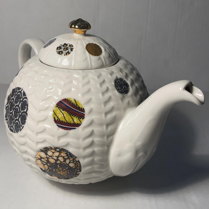 May include: White ceramic teapot with a textured surface and colorful, patterned circles. The lid is topped with a gold-colored knob. The spout and handle are smooth and white. A decorative tea accessory.