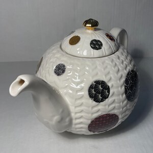 May include: White ceramic teapot with a textured surface and a lid adorned with black, gold, and burgundy circular patterns. The teapot features a gold-colored knob and a curved spout, perfect for serving tea.