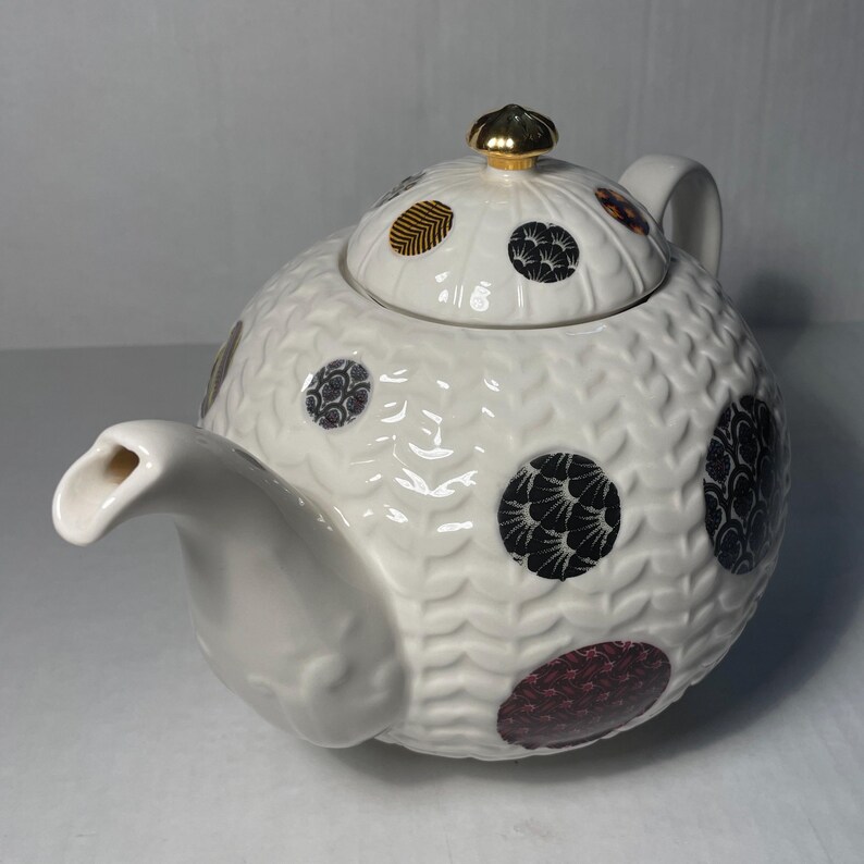 May include: White ceramic teapot with a textured surface and a lid adorned with black, gold, and burgundy circular patterns. The teapot features a gold-colored knob and a curved spout, perfect for serving tea.