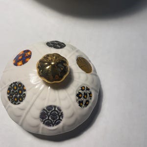 May include: A white ceramic lid with a gold knob and patterned circular designs in blue, yellow, and black. The lid has a scalloped edge and is likely part of a decorative container or serving dish.