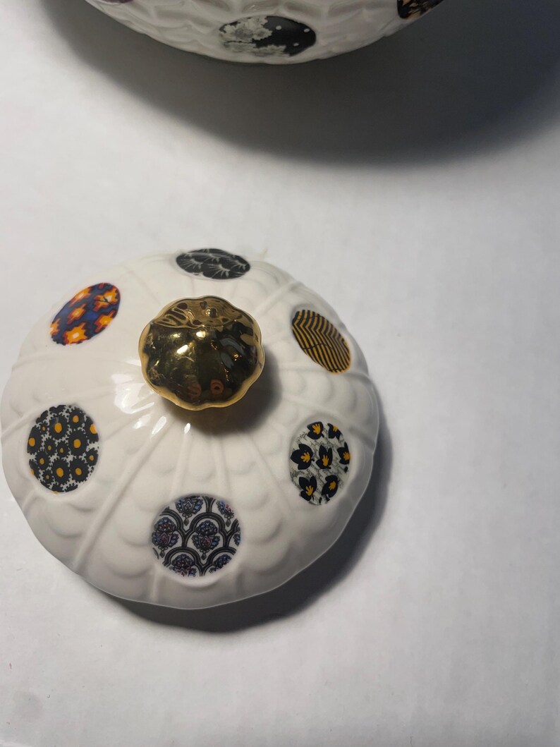 May include: A white ceramic lid with a gold knob and patterned circular designs in blue, yellow, and black. The lid has a scalloped edge and is likely part of a decorative container or serving dish.