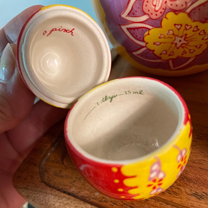 May include: A small, round, ceramic measuring container with a lid, featuring a vibrant yellow, red, and purple floral pattern. The interior of the lid is inscribed with the text a pinch, and the interior of the base reads 1 tbsp - 15 ml.