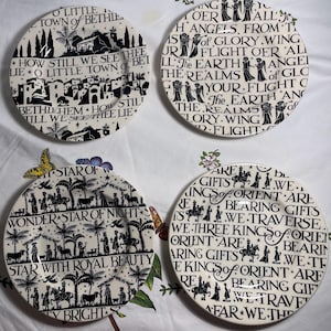 May include: Four round ceramic plates featuring black and white illustrations and text. The designs depict nativity scenes, angels, and the three kings with lyrics from Christmas carols including O Little Town of Bethlehem and We Three Kings.