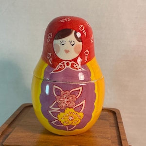 May include: A ceramic nesting doll featuring a red head covering with white floral outlines, a painted face with closed eyes, and a purple apron section decorated with large pink and yellow flowers against a bright yellow body base.