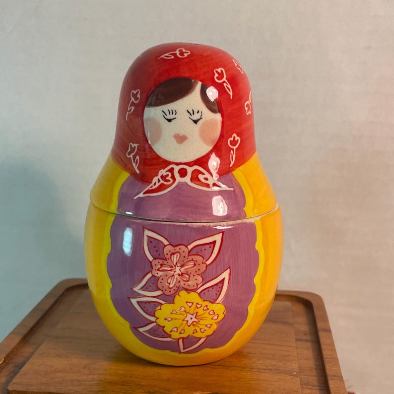 May include: A ceramic nesting doll featuring a red head covering with white floral outlines, a painted face with closed eyes, and a purple apron section decorated with large pink and yellow flowers against a bright yellow body base.