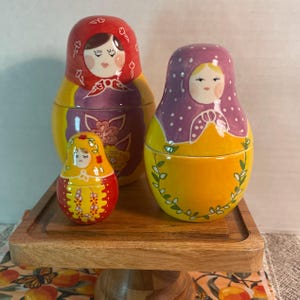 May include: Three nesting dolls with painted faces and colorful clothing. The largest doll is red and yellow with floral patterns. The medium doll is purple and yellow with white polka dots. The smallest doll is red and yellow with floral details.