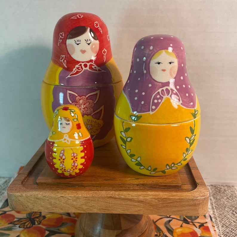 May include: Three nesting dolls with painted faces and colorful clothing. The largest doll is red and yellow with floral patterns. The medium doll is purple and yellow with white polka dots. The smallest doll is red and yellow with floral details.