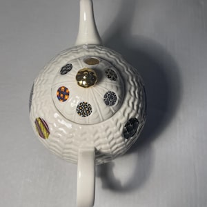 May include: White ceramic teapot with a textured surface and a gold-colored knob on the lid. The teapot is decorated with colorful, patterned circles. The handle and spout are white.