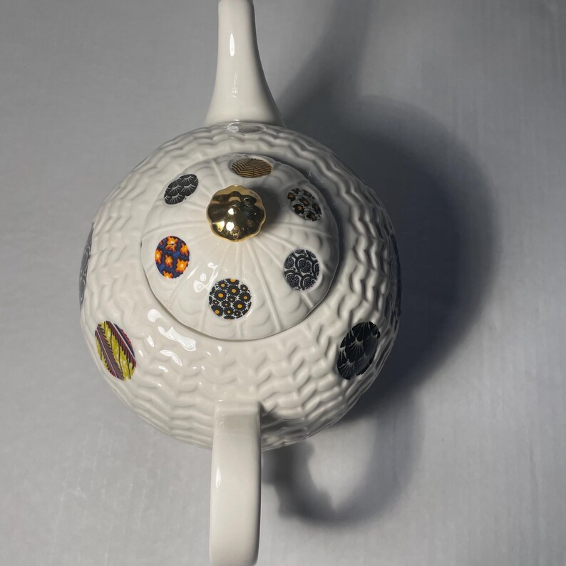 May include: White ceramic teapot with a textured surface and a gold-colored knob on the lid. The teapot is decorated with colorful, patterned circles. The handle and spout are white.