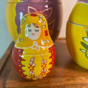 May include: A small, glossy, ceramic nesting doll featuring a yellow head covering with painted green vines and red flowers. The doll has a painted face with closed eyes and a red body decorated with yellow and purple floral patterns and dots.