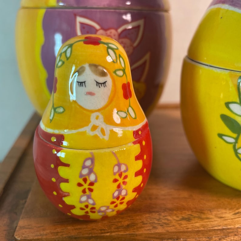 May include: A small, glossy, ceramic nesting doll featuring a yellow head covering with painted green vines and red flowers. The doll has a painted face with closed eyes and a red body decorated with yellow and purple floral patterns and dots.