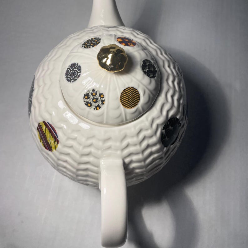 May include: White ceramic teapot with a textured, wavy pattern. The lid features a gold knob and is decorated with colorful, patterned circles. The teapot has a curved handle and a spout, perfect for serving tea.