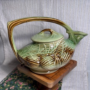 May include: A ceramic teapot with a green and brown leaf design. The teapot has a large handle and a lid with a small knob. The teapot is sitting on a wooden stand.